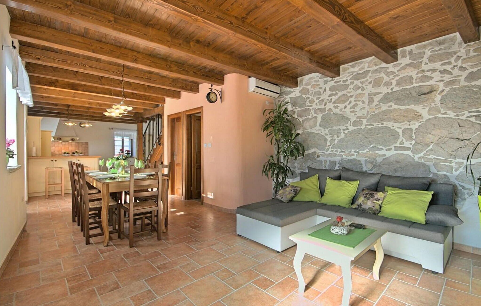 A spacious living area with a stone wall, wooden ceiling beams, a dining table with chairs, and a sofa with cushions.