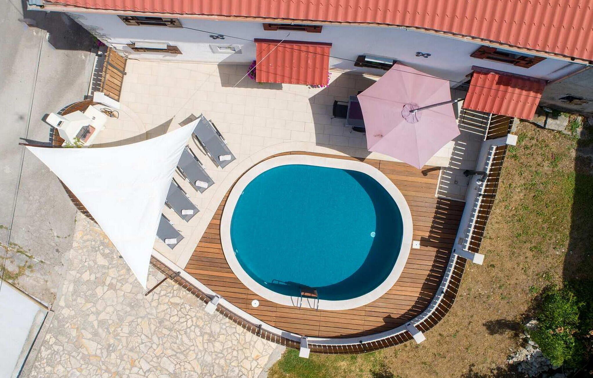 Vista aérea de una propiedad con piscina rodeada por una terraza de madera, un área de estar cubierta y un edificio con techo rojo.