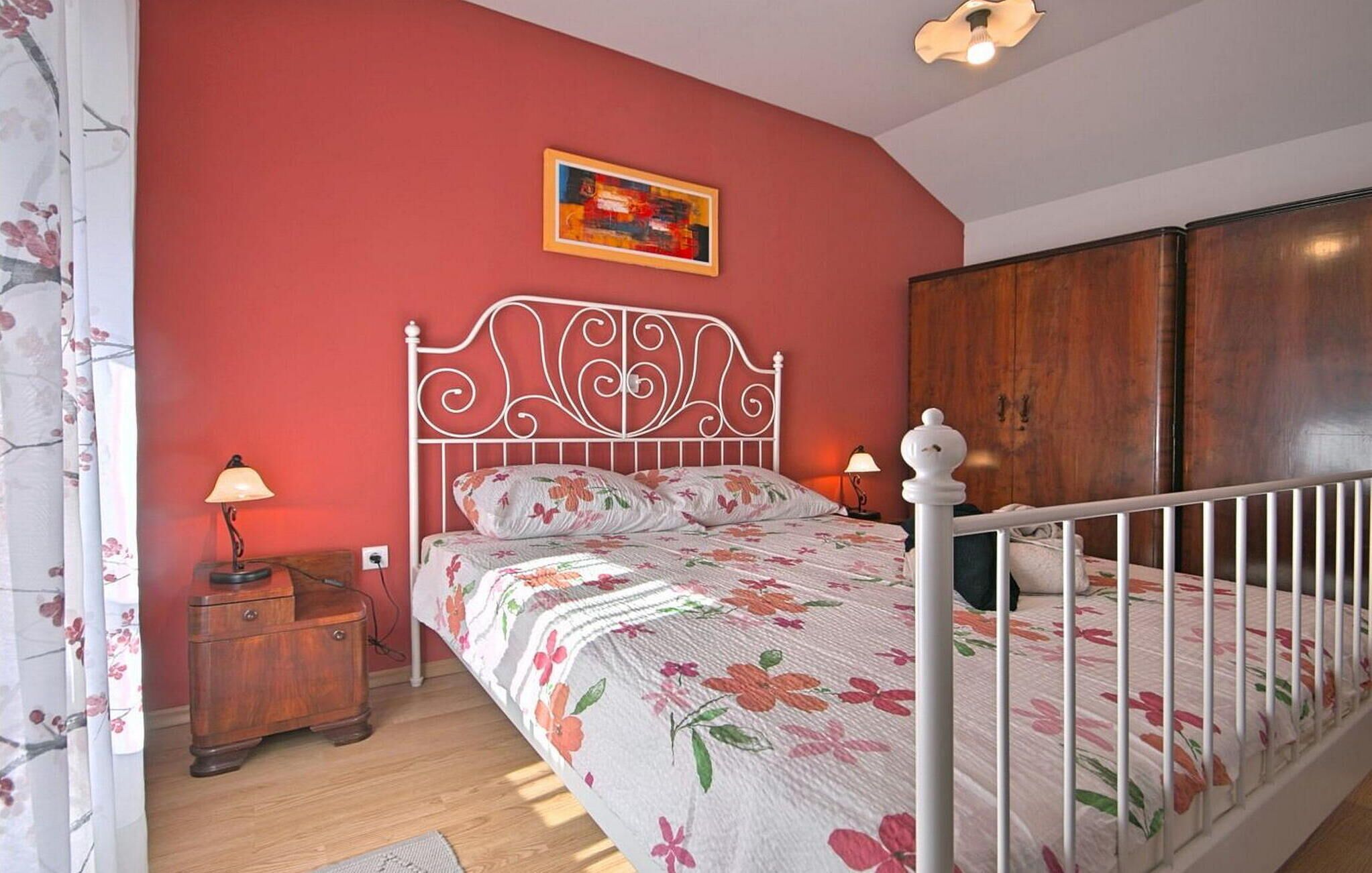 A bedroom with a floral bedspread, a white metal headboard, a wooden wardrobe, and a nightstand with a lamp.
