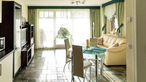 A modern living room with a glass dining table, a sofa, and a kitchen area with a TV and cabinets.