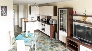 A compact kitchen with white cabinets, a dining area with a glass table, and a TV on a wooden stand. - Mühlenstr. 19 Wg.2, Deutschland (Zingst)