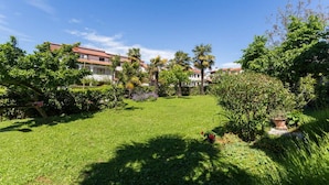 Un jardí ben cuidat amb una varietat de plantes i arbres, un cel blau clar i un edifici amb teulades de color vermell al fons.