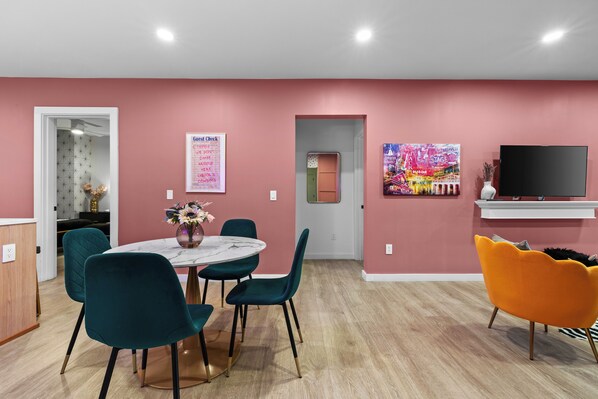 A modern living room with a pink wall, a round table with chairs, a TV, and artwork.