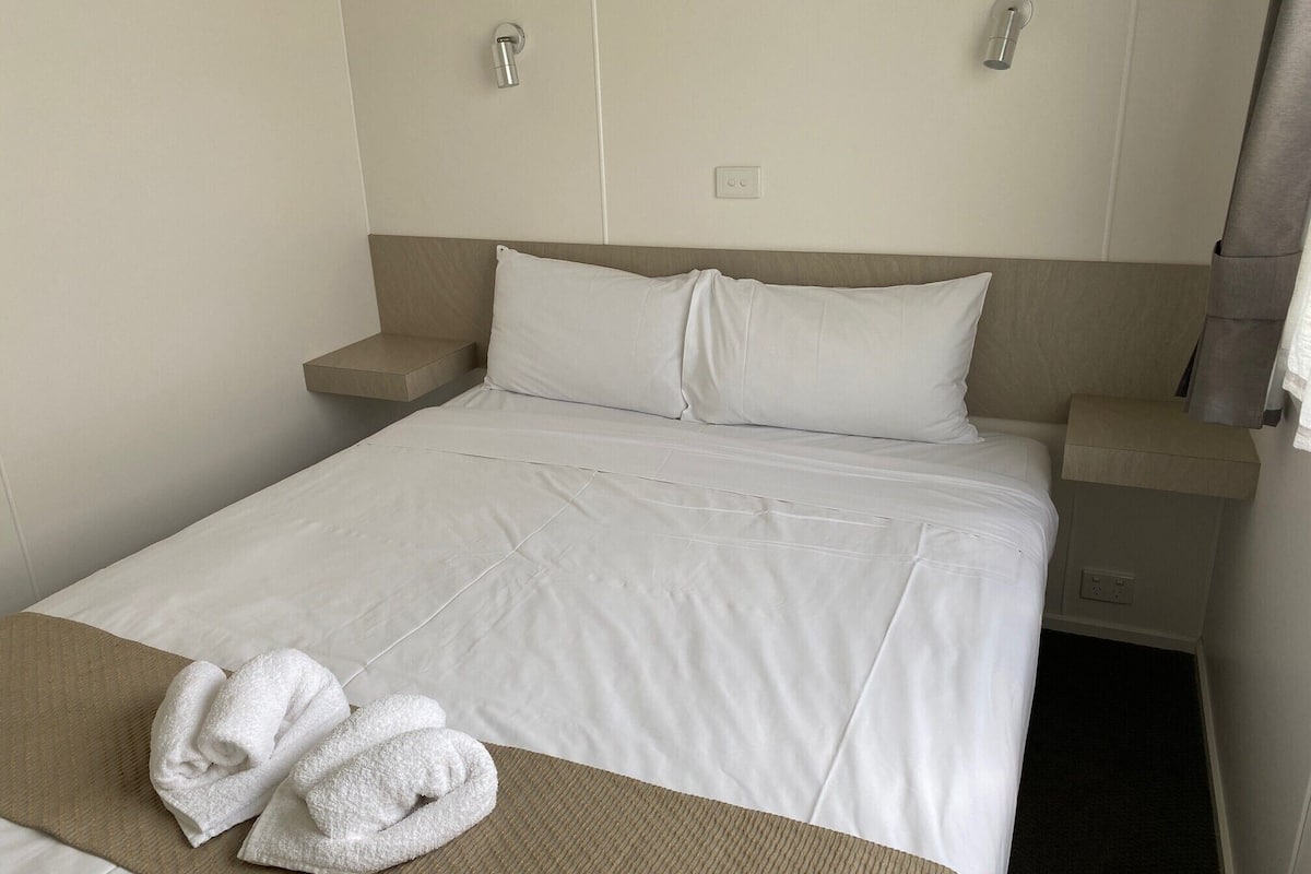 Una cama bien tendida con sábanas blancas y dos almohadas, una manta marrón y dos toallas blancas dobladas sobre la cama.