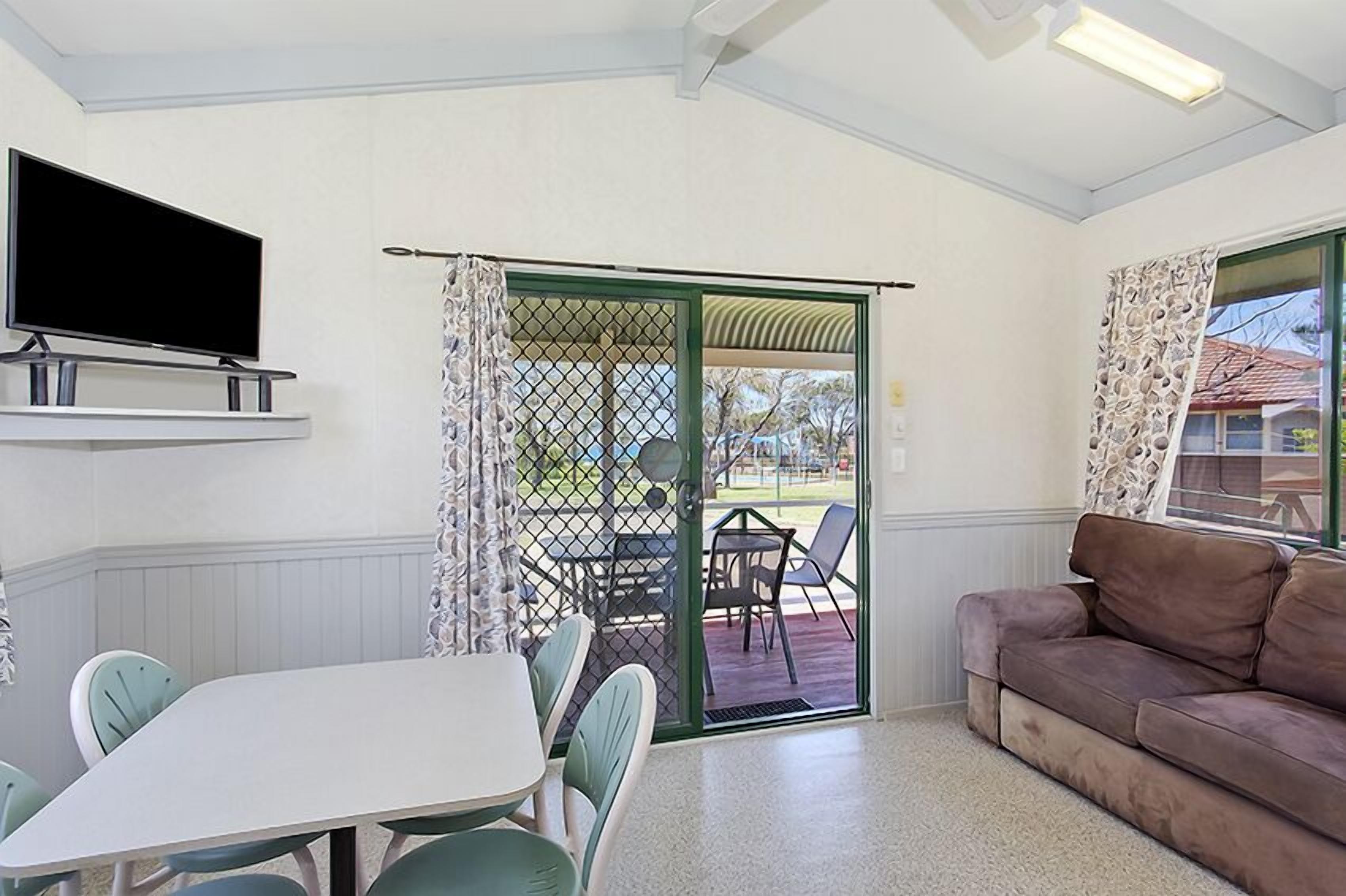Una habitación con una televisión montada en la pared, una mesa de comedor con sillas, un sofá y un ventanal corredero que da a un área exterior con una mesa y sillas.