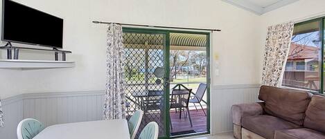 A room with a television mounted on the wall, a dining table with chairs, a sofa, and a sliding glass door leading to an outdoor area with a table and chairs.