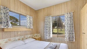 Un dormitorio con un muro revestido de madera, una cama con ropa blanca, dos ventanas con cortinas estampadas y vistas a la vegetación exterior.