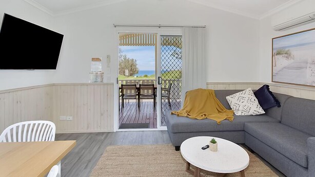 Un salón con un sofá gris, una mesa de centro blanca, un televisor montado en la pared y un ventanal corredizo que da a un patio exterior con una mesa y sillas.