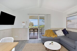 Un salon avec un canapé gris, une table basse blanche, un téléviseur fixé au mur et une porte-fenêtre coulissante donnant sur une terrasse extérieure avec une table et des chaises.