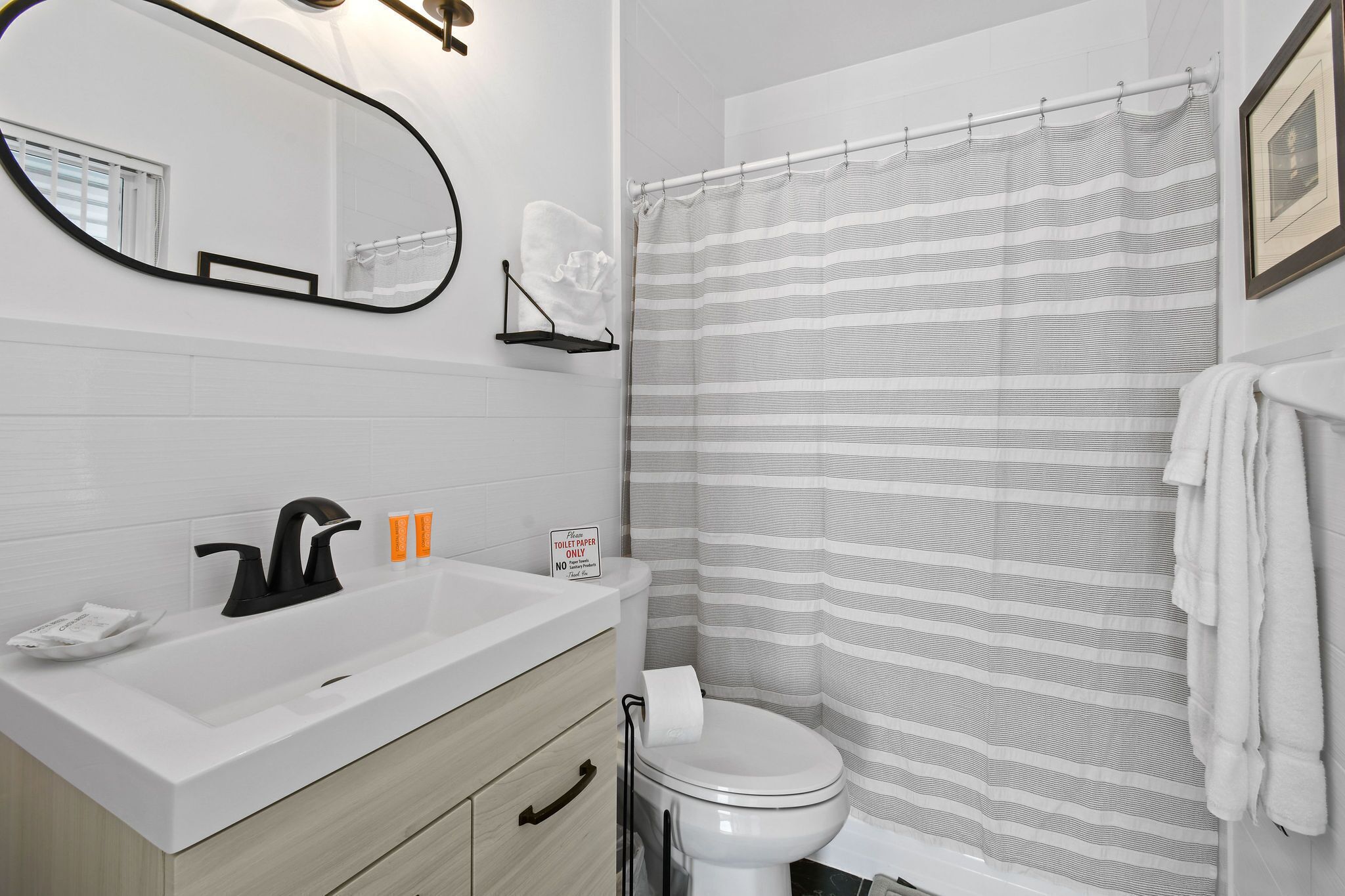 Une salle de bain avec un lavabo blanc, un robinet noir et des toilettes. On y trouve un rideau de douche à rayures et une serviette accrochée au mur.