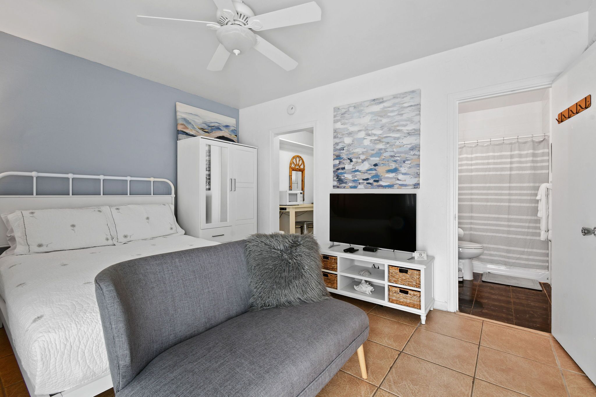Une chambre à coucher avec un lit, un canapé gris, un téléviseur posé sur un meuble et une salle de bain visible à travers une porte ouverte.