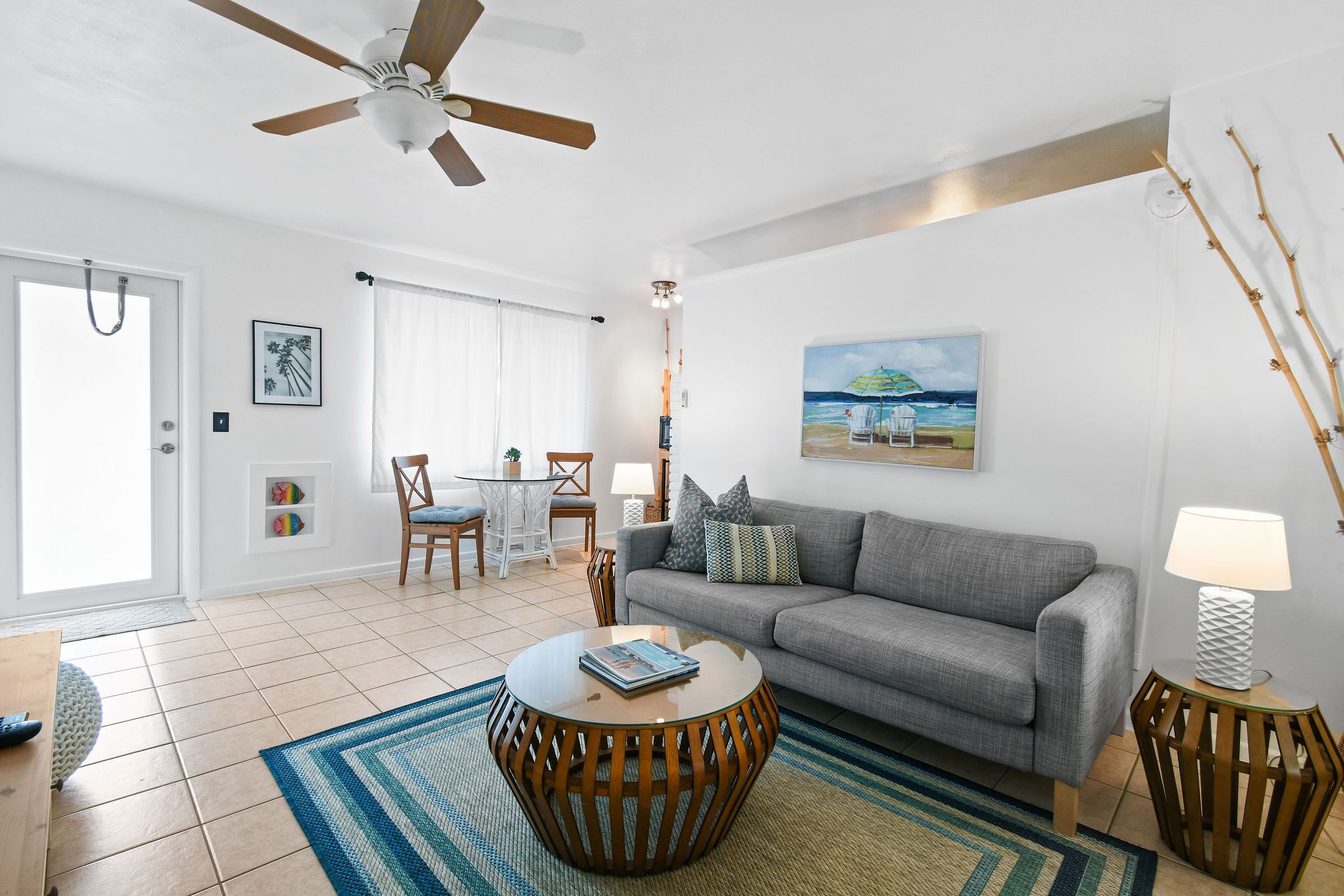 A living room with a grey sofa, round wooden coffee table, and a ceiling fan. There is a painting of a beach scene on the wall, a lamp on a side table, and a door with a towel hanging on it.
