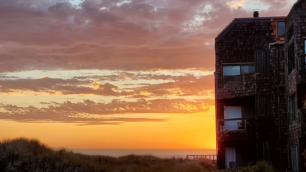 Un bâtiment moderne doté d’un toit plat et de nombreuses fenêtres, situé près d’une étendue d’eau, avec un coucher de soleil en arrière-plan.