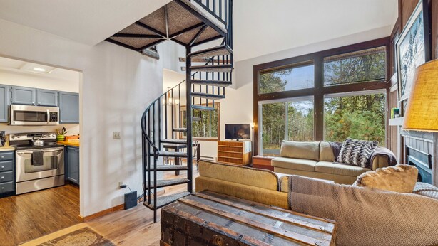 Una cocina moderna con electrodomésticos de acero inoxidable, piso de madera y una escalera espiral que lleva a un dormitorio estilo loft con un amplio ventanal con vistas a árboles.