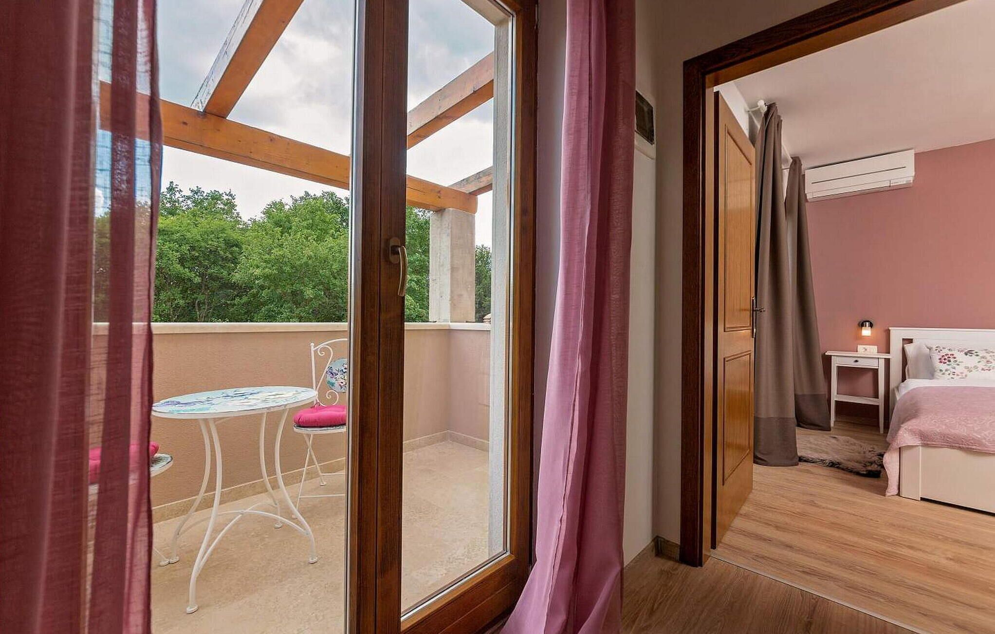 A room with a bed, a bedside table, and a window overlooking a balcony with a table and chairs.