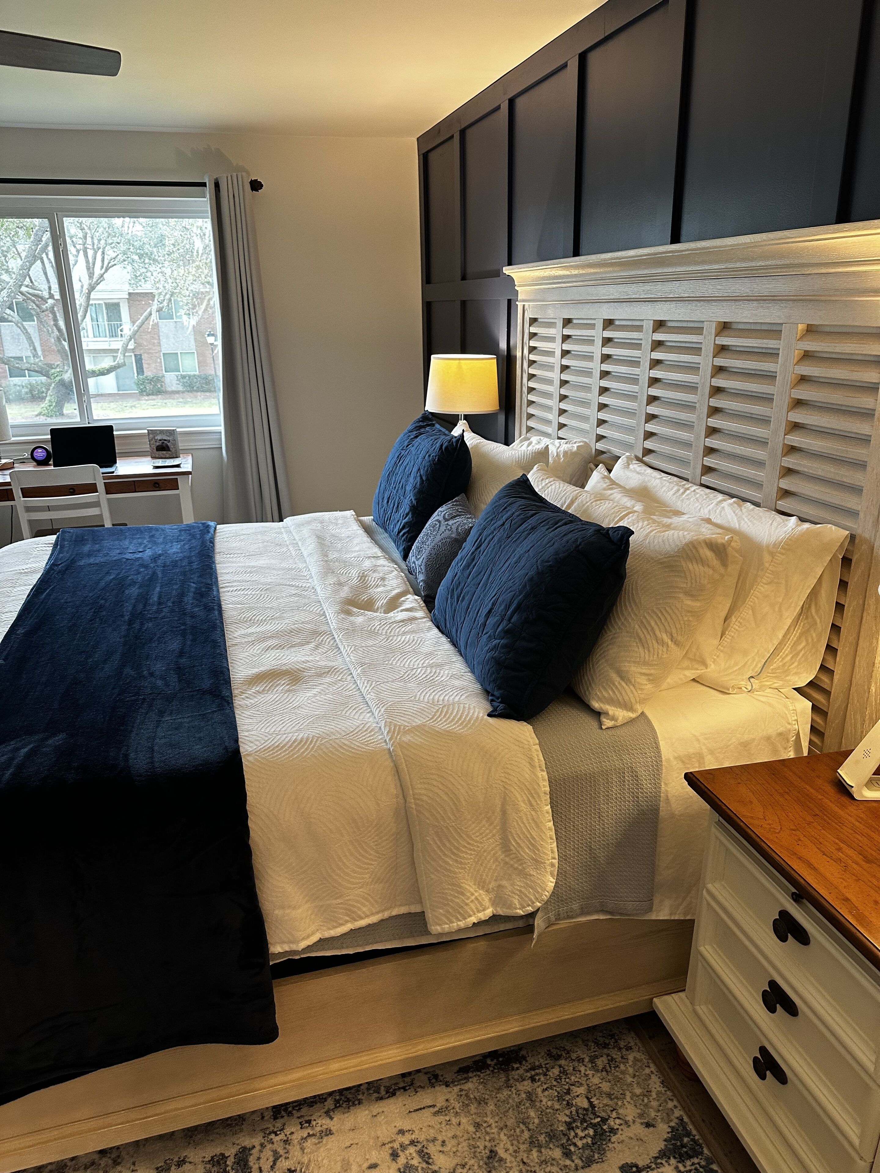 A neatly made bed with a blue throw blanket, a bedside table with a lamp, and a view of the outdoors through a window.