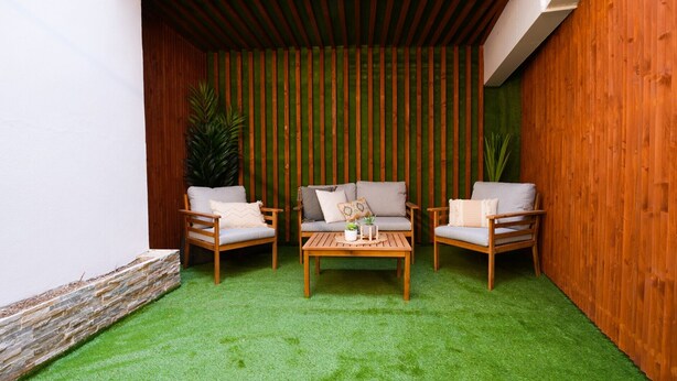 Een klein terras met twee houten stoelen en een tafel, omringd door een houten muur en een vloer van groen kunstgras.