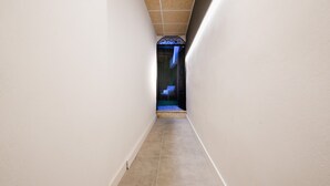 Een smalle gang met witte muren en een betegelde vloer, leidend naar een kamer met een blauw verlichte deuropening.