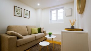 Een woonkamer met een beige bank, een kleine ronde tafel, een potplant en ingelijste botanische prints aan de muur.
