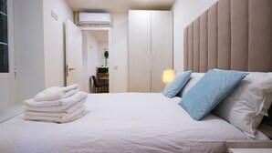En pænt redt seng med hvide sengetæppe og et lag hvide håndklæder, en sengetavle med lodrette paneler og en bordlampe.