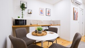 Et kompakt køkken med mikroovn, vask og lille bordplade. To indrammede kunstværker på væggen og en potteplante hængende over bordet.