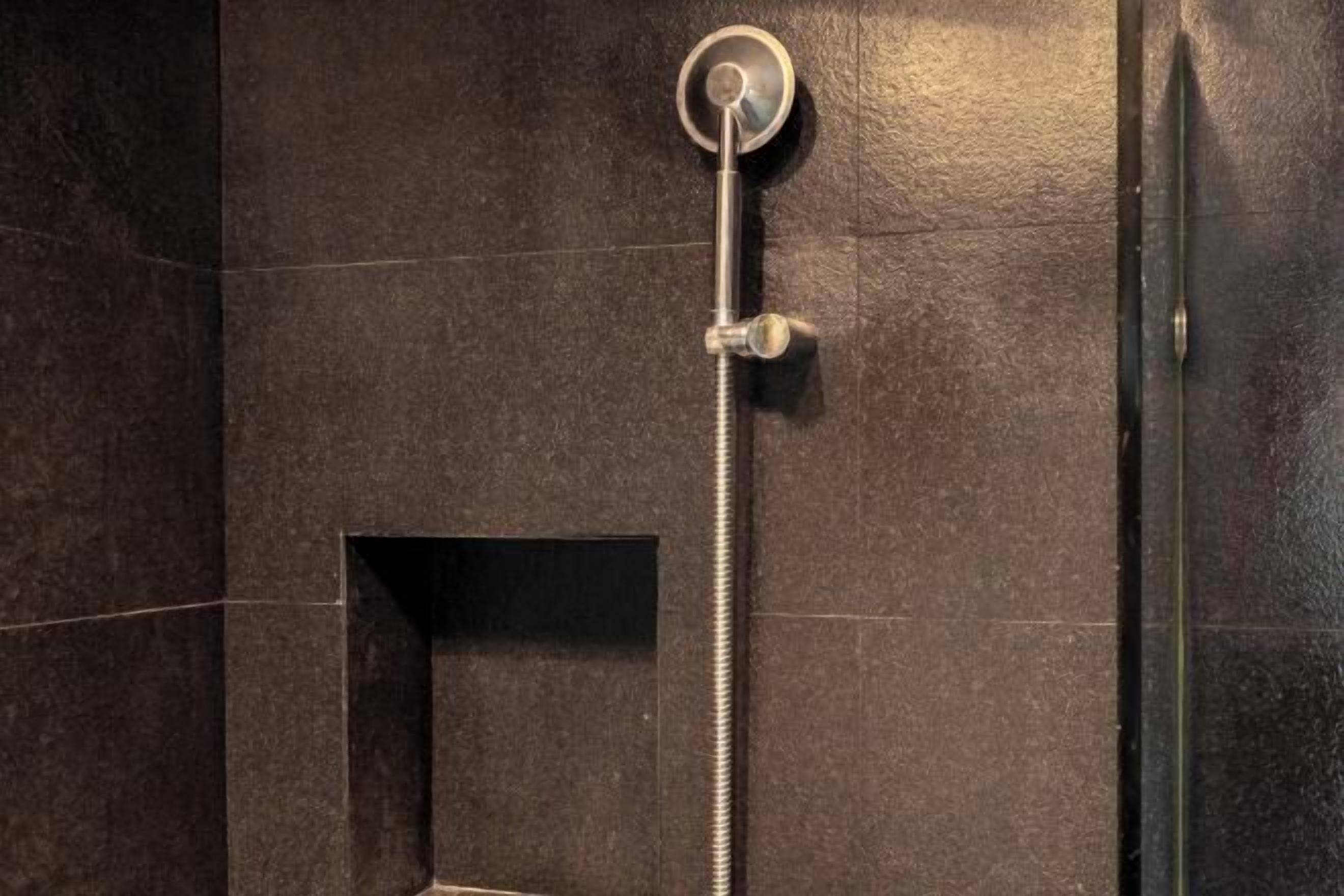A shower with a handheld showerhead mounted on a wall, a control panel, and a rectangular opening in the wall.