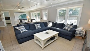 A spacious living room with a sectional sofa, a white coffee table, and large windows offering views of the outdoors. - Game Time - Channel Front in Garden City Beach (Garden City Beach)