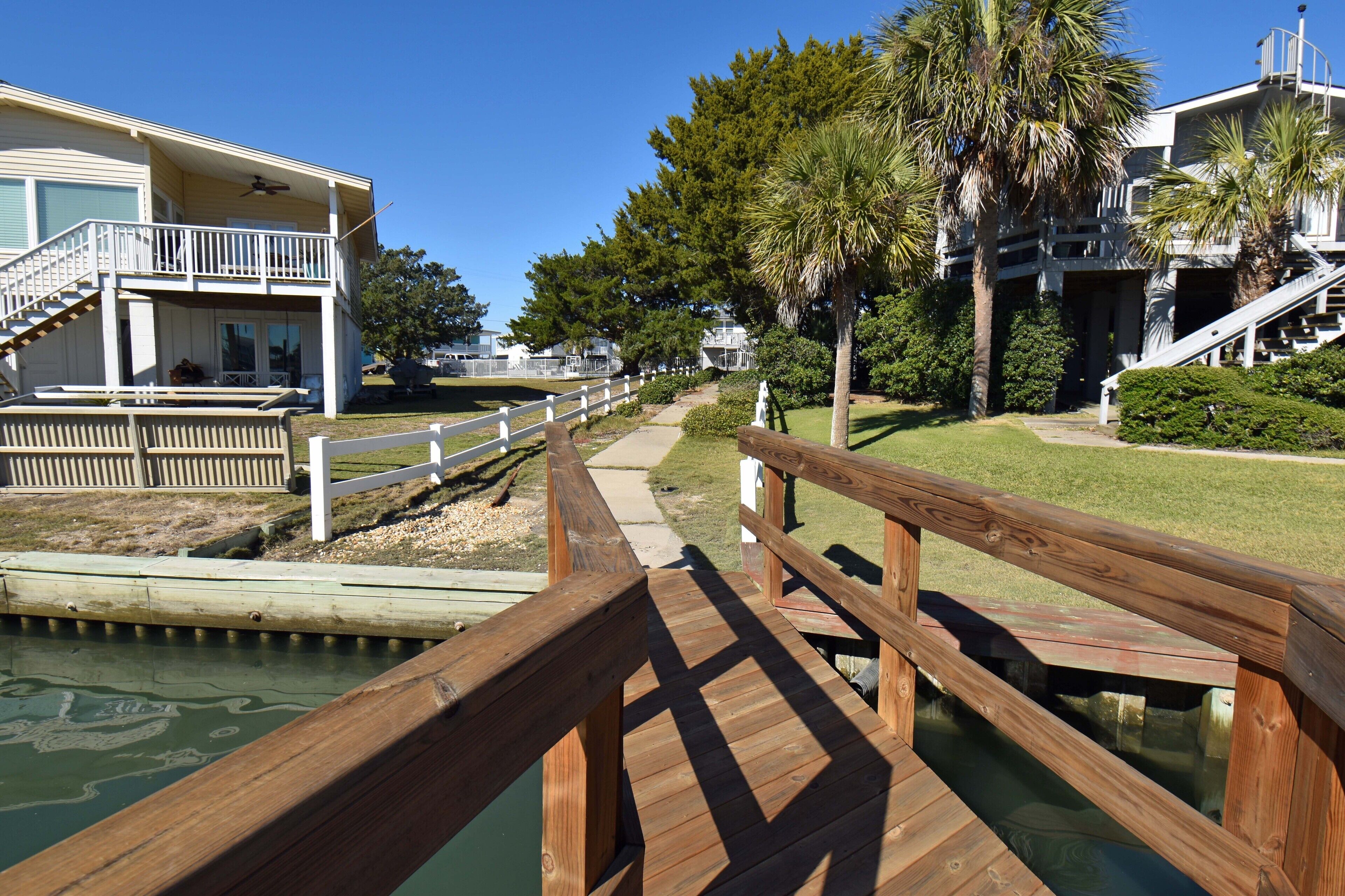 A wooden boardwalk leading to a building with a balcony, surrounded by greenery and a clear sky.