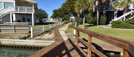 A wooden boardwalk leading to a building with a balcony, surrounded by greenery and a clear sky.