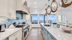A modern kitchen with a view to the sea, featuring white cabinetry, a blue backsplash, and a large island with a bowl. - Jonesin' By The Beach - Ocean Front & Channel Front in Garden City Beach (Garden City Beach)