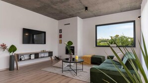 A modern living room with a flat-screen TV, a green sofa, a coffee table, and a large window offering a view of greenery and a building. - Amazing home in Visnjan (Visnjan)