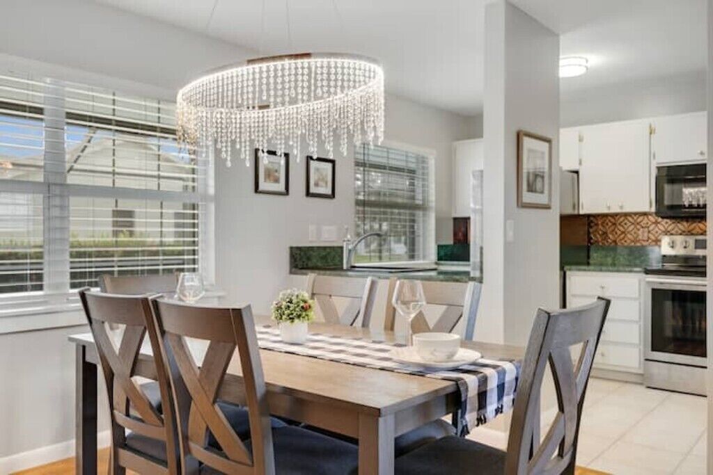Un coin repas avec une table en bois, des chaises et un lustre. On y trouve des fenêtres avec des stores, une cuisine à l’arrière-plan et des tableaux encadrés au mur.