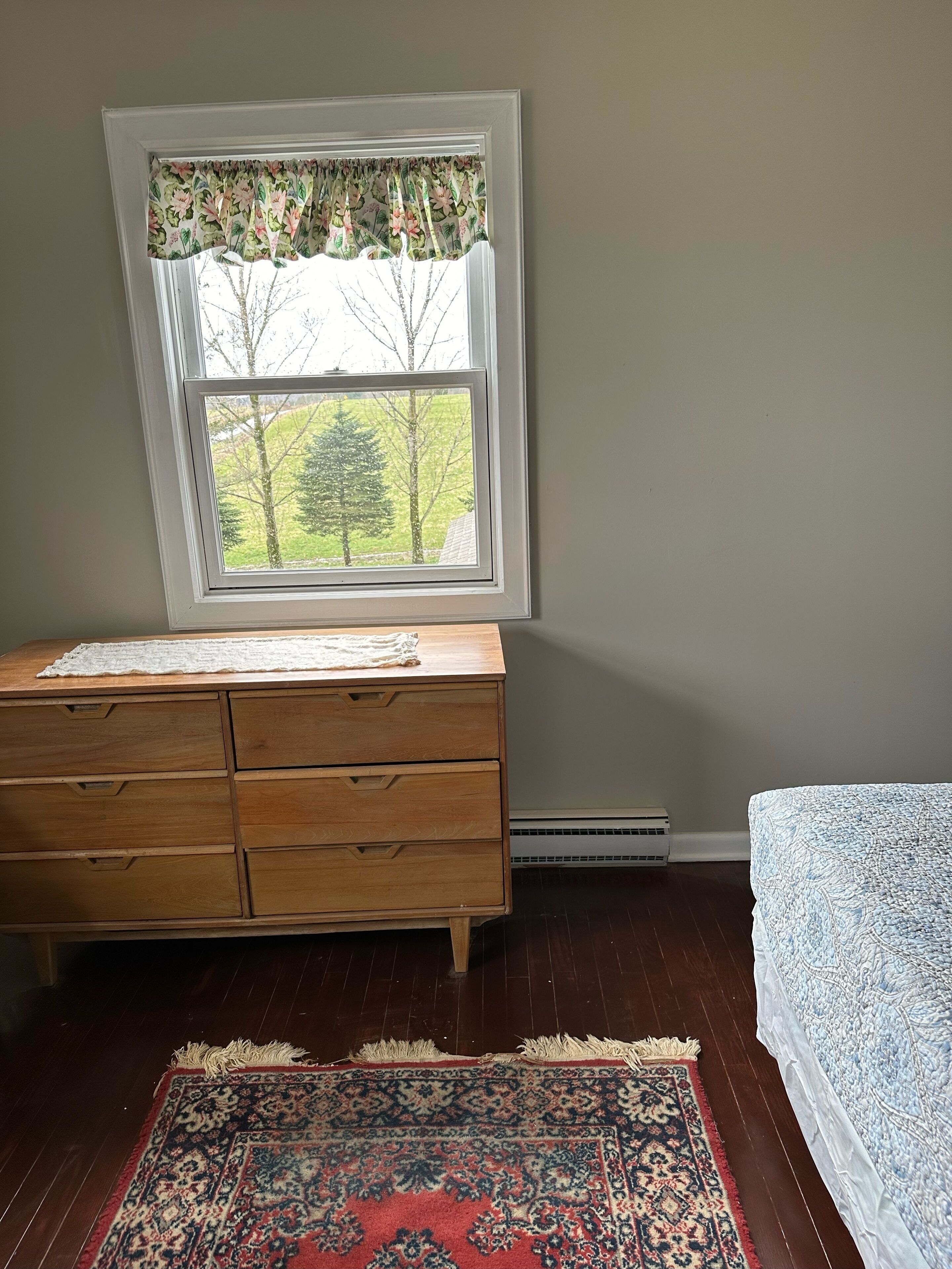 Una habitación con un aparador de madera, una ventana con cortinas florales, un tapete estampado y una cama con colcha azul y blanca.