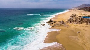 Una playa costera con aguas turquesas cristalinas, orilla arenosa, afloramientos rocosos y un edificio de resort al fondo.
