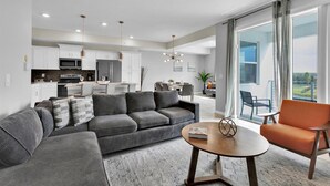Un salon moderne avec un canapé d’angle gris, une table basse ronde et un fauteuil orange. On aperçoit à l’arrière-plan une cuisine avec des meubles blancs et un coin repas comprenant une table et des chaises.