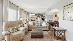 Amplia sala de estar con un sofá color beige, un reposapiés marrón y un comedor con mesa y sillas de madera.