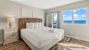 Un dormitorio con una cama grande, una mesita de noche con lámpara, vista al océano a través de una ventana, y un balcón con una silla.