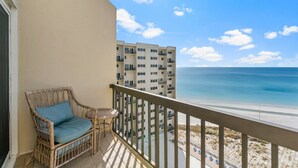 Un balcón con una silla de mimbre y una mesita pequeña, con vistas a la playa y un edificio de gran altura.
