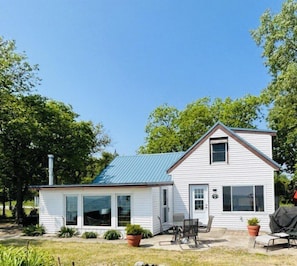 A two-story house with a blue roof, a covered patio with a table and chairs, and a small garden area with potted plants. - East Shore - Lake View Cottage - Pelee Island (Pelee)