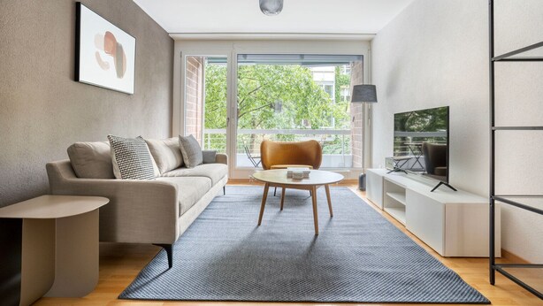 Una sala moderna con un sofá, una mesa de centro, un televisor sobre un mueble bajo y un amplio ventanal con vistas a la vegetación.