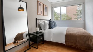 Un dormitorio moderno con un espejo grande, una cama con edredón blanco, un escritorio de madera con una lámpara y vistas a un edificio de ladrillo a través de una ventana.