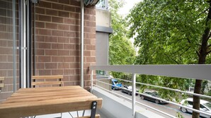 Un balcón con una mesa y sillas de madera, barandilla metálica y vista a coches estacionados y árboles.