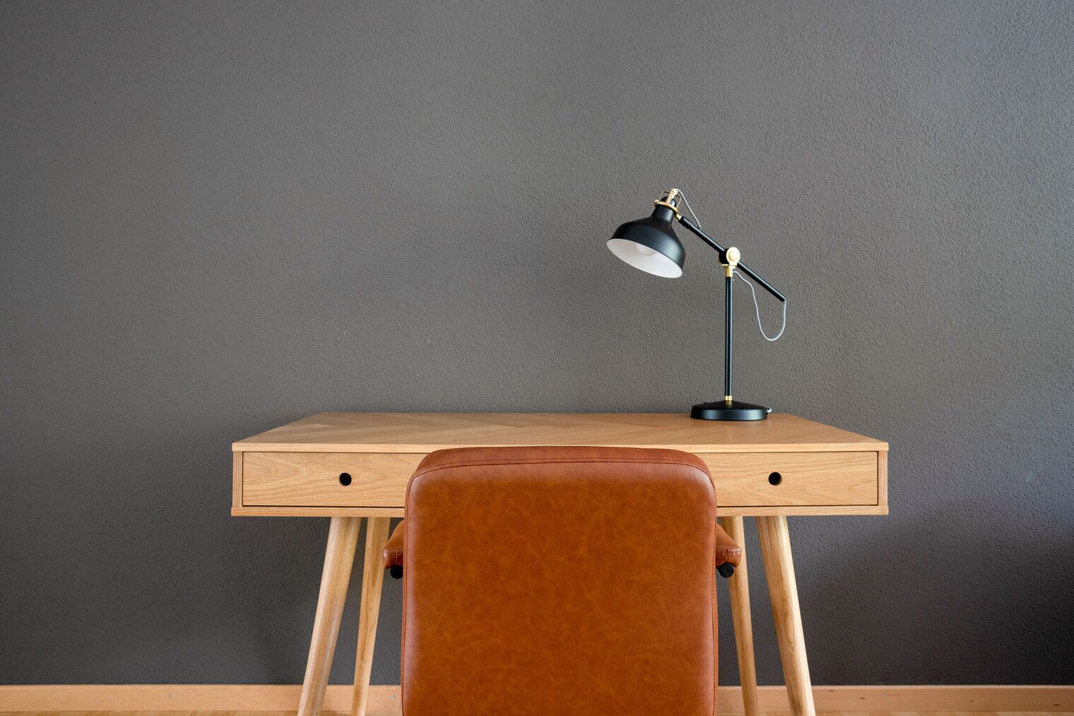 A wooden desk with a black desk lamp, a brown leather chair, and a grey wall.