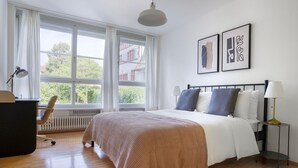 A bedroom with a large bed, a desk, and a chair. There are two windows with curtains, a radiator, and a wall-mounted lamp.