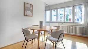 A modern dining area with a round wooden table and four chairs, a framed abstract artwork on the wall, and a large window with a view of buildings.