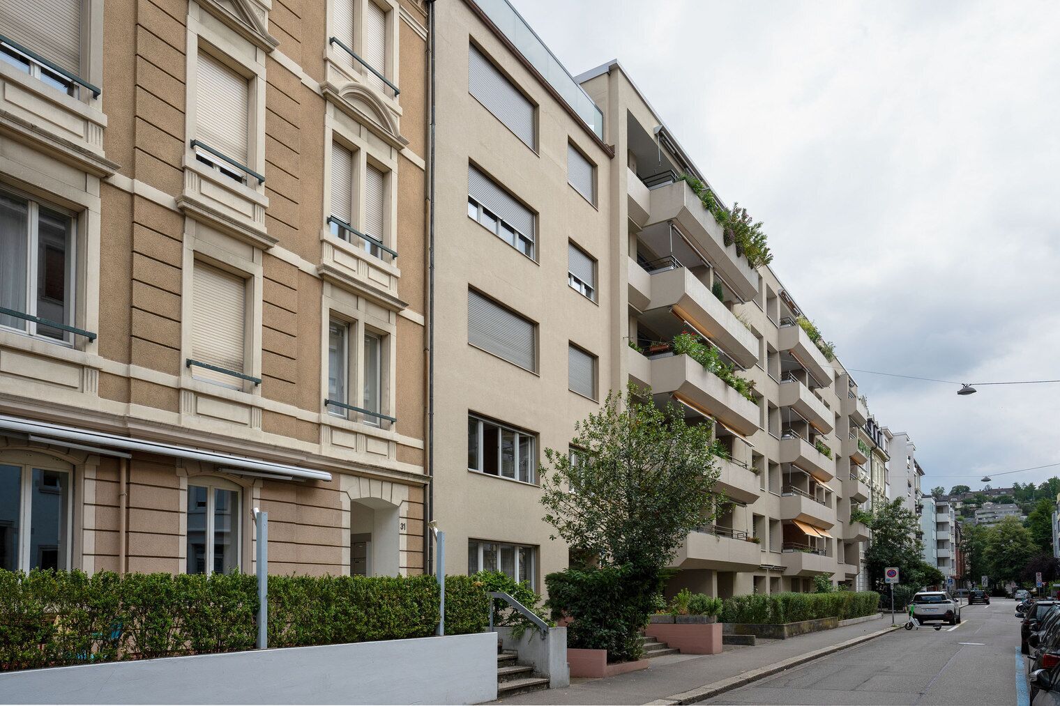 A multi-story residential building with balconies, a tree in the foreground, and a street with parked cars.