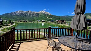 Un pontile in legno con tavolo e sedie in metallo affacciato su un lago, circondato da montagne e case.