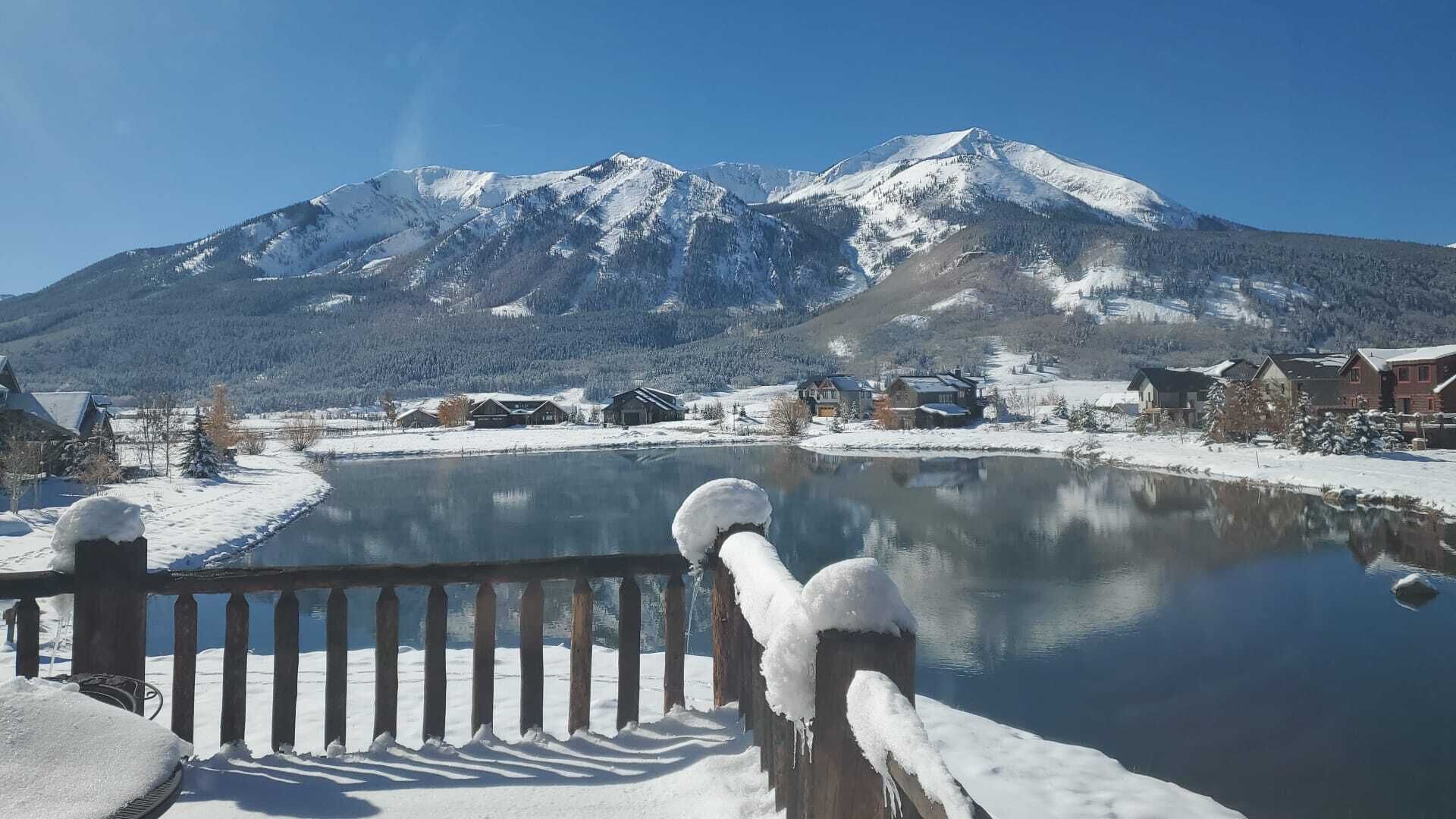 A snow-covered landscape with a mountain in the background, a wooden fence in the foreground, and a body of water reflecting the scenery.