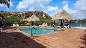 Un espace piscine doté de chaises longues et de parasols en chaume, entouré d’une colline rocheuse et d’une étendue d’eau.