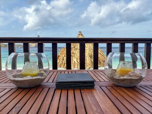 Duas taças com bebidas e conchas do mar sobre uma mesa de madeira, com vista para o oceano e uma construção de palha.